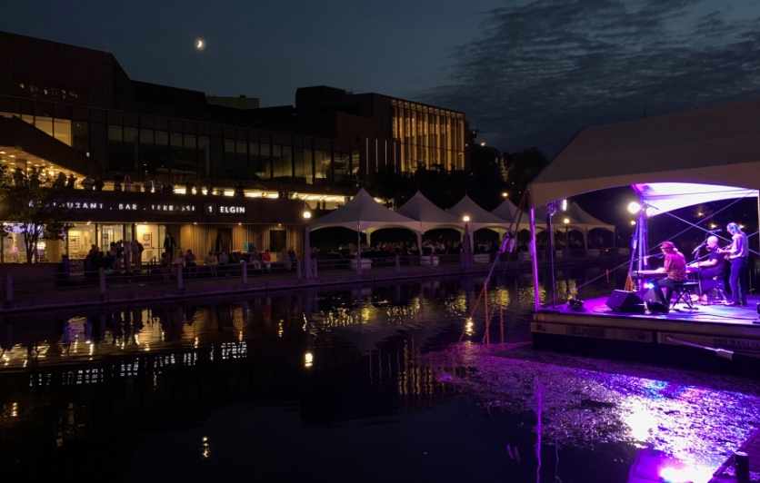 View of a band playing from a stage floating on the Rideau Canal to diners on the 1 Elgin patio, at night. 